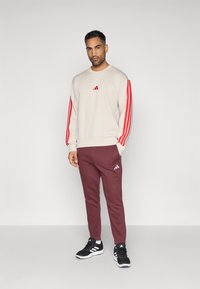 Egy férfi bézs pulóvert visel, melynek ujjain piros csíkok találhatók, bordó melegítőt és fekete sportcipőt, amin fehér csíkok vannak.