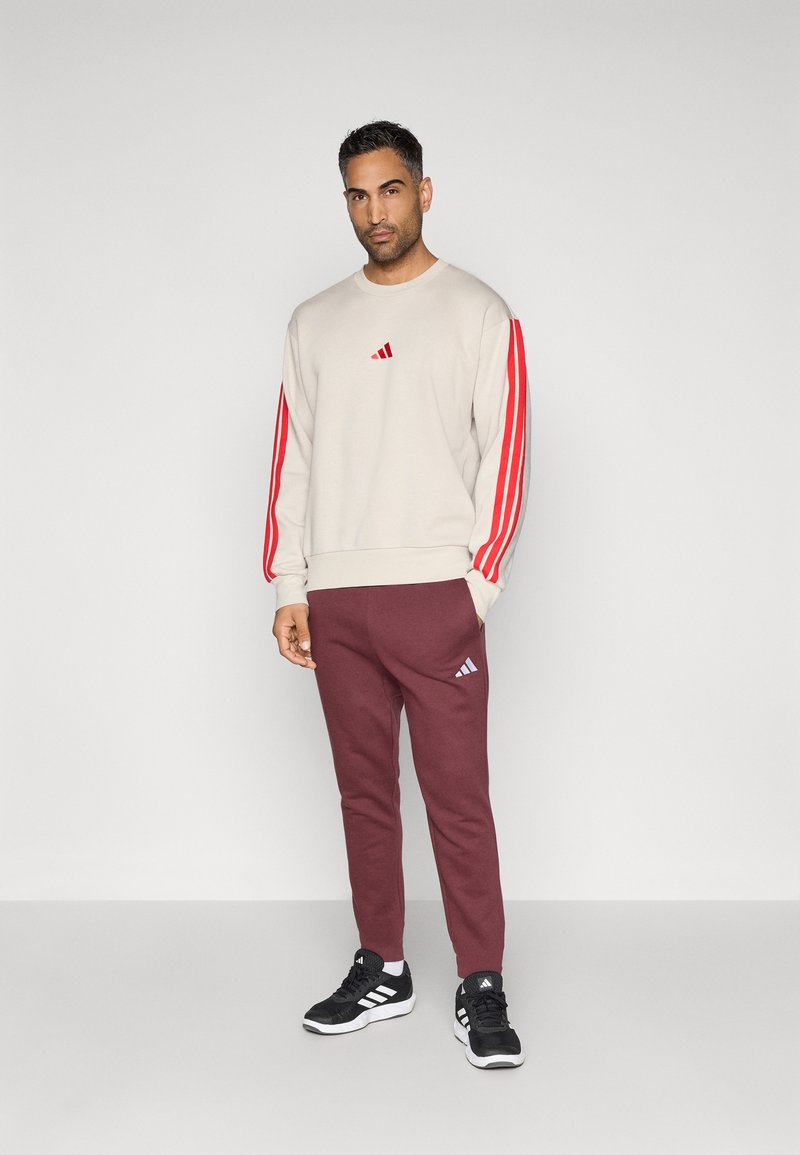 Man staat in een beige sweatshirt met rode strepen op de mouwen, bordeauxrode joggingsbroek en zwarte sneakers met witte strepen.