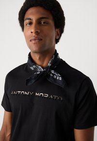 Tricou negru cu logo metalic "ANTONY MORATO" și o bandană neagră cu modele paisley albe legată în jurul gâtului.