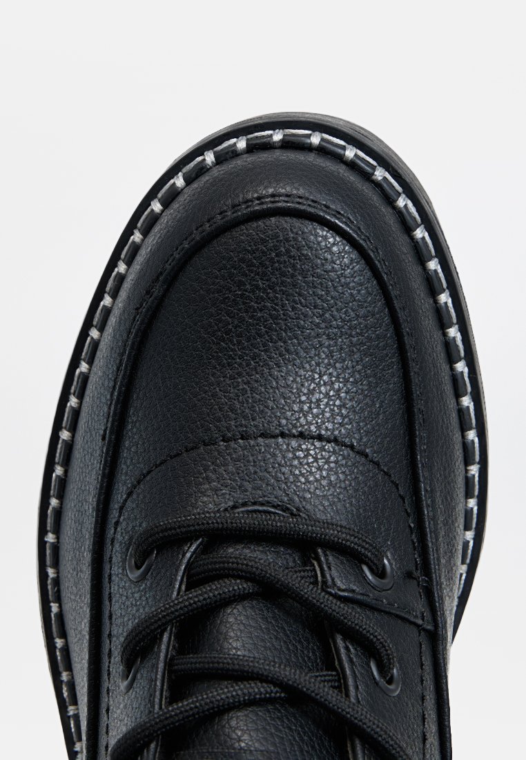 Zapato de cuero negro con punta redonda, acabado texturizado, detalle de costura blanca a lo largo de la suela y cordones entrelazados.