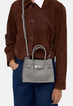 Osoba ubrana w ciemnobrązową zamszową kurtkę i niebieskie dżinsy trzyma szarą torebkę o teksturze krokodyla z górnymi uchwytami i regulowanym paskiem na ramię.