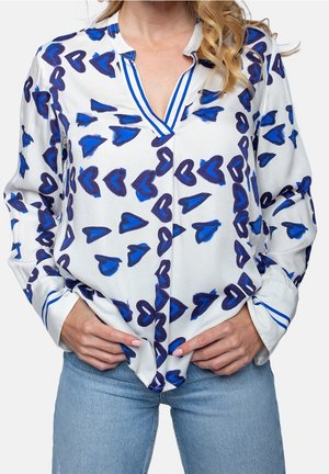 Blouse blanche avec des motifs de cœurs bleus, col et poignets rayés, portée rentrée dans un jean bleu clair, mains tenant l'ourlet de la chemise.