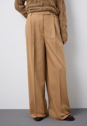 Persona con pantalones anchos beige, un suéter de punto marrón y zapatos planos oscuros, de pie contra una pared blanca lisa.