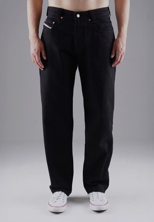 Man wearing black straight-leg jeans and white sneakers standing against a plain gray background.