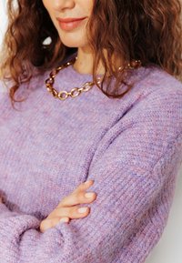 Femme aux cheveux bouclés portant un collier en chaîne dorée épaisse et un pull lavande texturé, bras croisés.