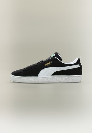 Sneaker Puma in suede nero con striscia laterale bianca, suola bianca e lacci neri, mostrata di profilo su sfondo neutro.