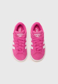 Pink suede sneakers featuring white stripes and eyelets, flat laces, and a round toe design with a textured sole.