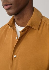 Camisa de pana color mostaza con un cuello estructurado y botones blancos, que presenta una sutil textura acanalada y está superpuesta a una camiseta blanca.