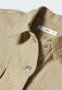 Chemise beige à boutons avec un col structuré, des éléments en métal argenté et des détails de couture visibles. La texture du tissu est lisse et durable.