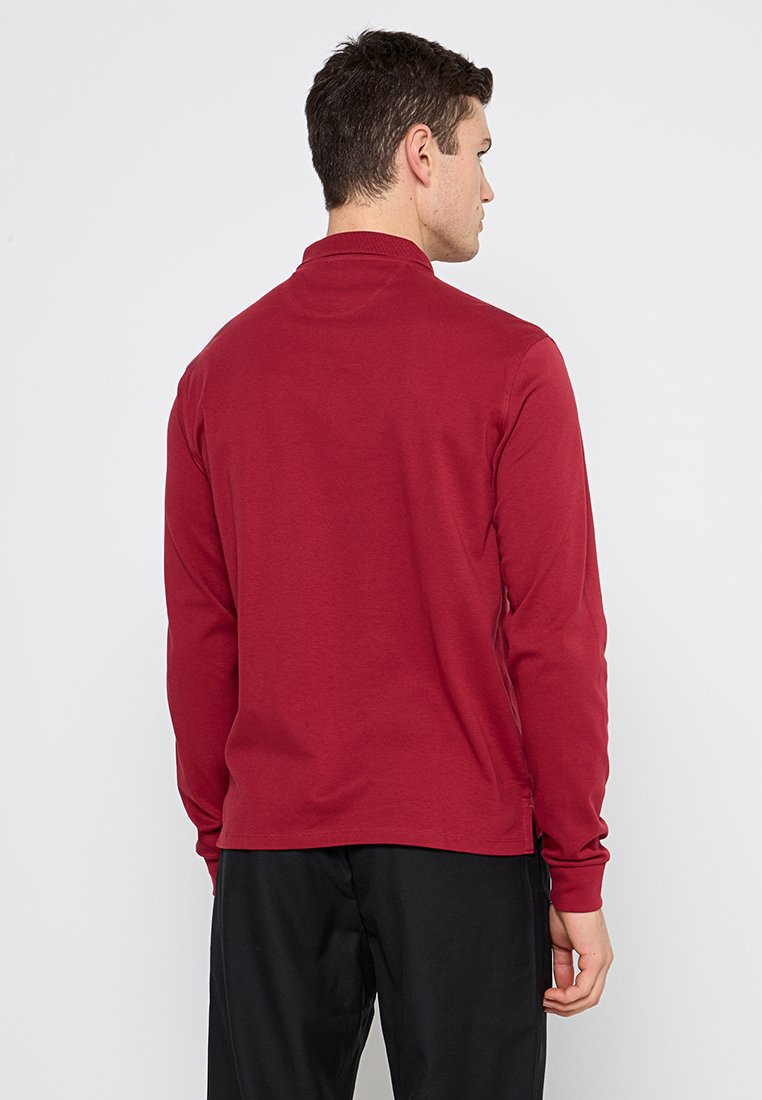 Chemise à manches longues de couleur rouge profond, en coton doux, avec un col rond et des coutures minimales, associée à un pantalon noir.