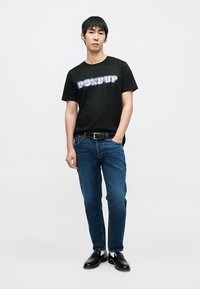 T-shirt noir avec inscription "DON DUP" en bleu et blanc, associé à un jean bleu et des chaussures noires, posé sur un fond uni.
