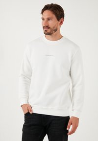 Biała bluza z miękkiego materiału, posiadająca długie rękawy i okrągły dekolt. Zawiera subtelną haftowaną aplikację logo w okolicy klatki piersiowej.