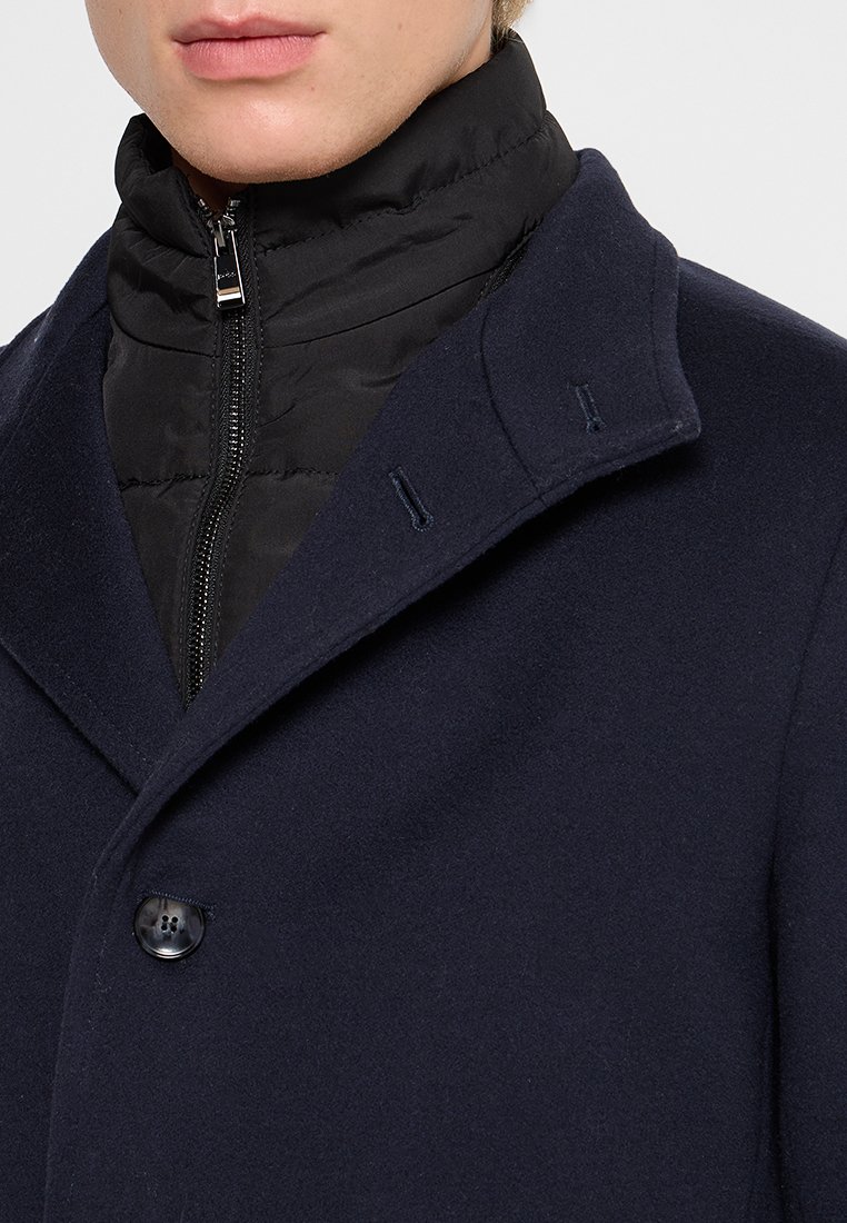Manteau en mélange de laine bleu marine avec un col montant, présentant une doublure intérieure matelassée noire et une fermeture à un bouton à l'avant.