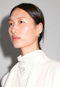 Model with pale skin and brown eyes wears a white, high-collared top. Notable features include a silver hoop earring and straight black hair.