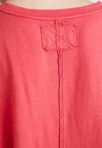Rood stof met een verticale naad en een rechthoekige patch die nabij het bovenste midden van de achterkant is gestikt, met gedetailleerde textuursteken.