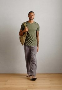 Oliven grønn kortermet t-skjorte, avslappede gråbukser og sandaler. Modell bærer en beige veske. Nøytral bakgrunn med treverk.