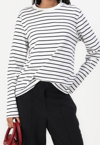 Persona che indossa una camicia bianca a maniche lunghe con strisce orizzontali nere e pantaloni neri, mentre tiene una borsa rossa al suo fianco.