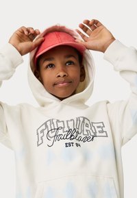 Sweat-shirt tie-dye blanc avec le texte noir "FUTURE Trailblazer", porté sous une casquette rose. Il possède une poche avant et des poignets côtelés.