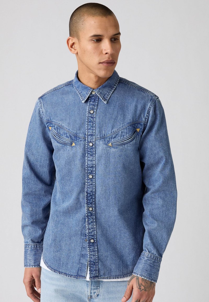 Lichtblauwe denim overhemd met een knoopsluiting aan de voorkant, puntige kraag, dubbele borstzakken en contrasterende pijl details op de zakken.