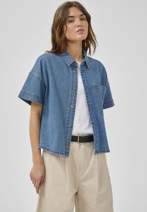 Vrouw draagt een loszittend denim overhemd met korte mouwen over een wit shirt, beige broek met hoge taille en een zwarte riem, staand tegen een effen achtergrond.