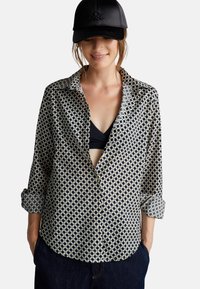 Lange mouwen knoopshirt in crème met een marineblauw geometrisch patroon, voorzien van een omgeslagen kraag en opgerolde mouwen, gecombineerd met een zwarte pet.
