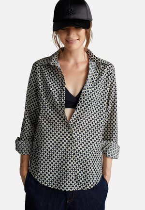 Chemise à manches longues boutons en crème avec un motif géométrique bleu marine, dotée d'un col rabattu et de poignets retroussés, accompagnée d'une casquette noire.