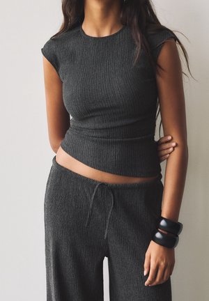Vrouw draagt een gestructureerd donkergrijs mouwloos topje en bijpassende losse broek met zwarte grove armbanden aan de rechterpols.