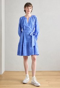 Rochie brodată de culoare albastră deschisă, cu mâneci lungi, decolteu în V, talie legată și fustă în straturi, asortată cu adidași albi tip high-top.