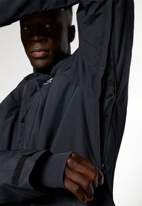 Jachetă neagră, waterproof, cu guler înalt, ventilație cu fermoar și manșete cu Velcro. Are un interior texturat și un design elegant, modern.