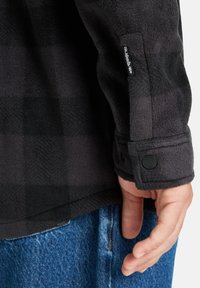 Camisa de franela a cuadros en negro y gris con textura de forro polar, puños abotonados y etiqueta de logo en la manga. Jeans de mezclilla visibles por debajo.