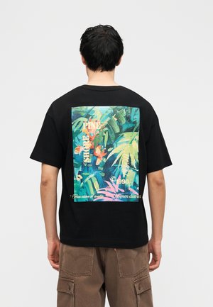 Personne portant un T-shirt noir avec un imprimé tropical coloré et du texte au dos, associé à un pantalon cargo marron.