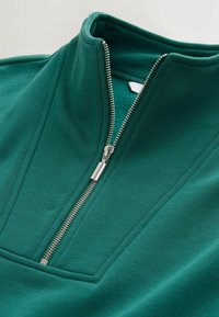 Sweat-shirt vert à fermeture quart de zip argentée partiellement ouverte, montrant l'intérieur doublé en polaire et le col relevé.