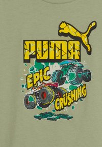 Gelbes, rissiges Puma-Logo über zwei bunten Monstertrucks, die drei grüne Autos auf einem olivgrünen Hintergrund zerquetschen. Die Worte "Episches Zerdrücken" in fettem Gelb.