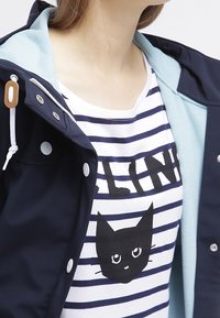 Chaqueta azul marino con forro azul claro, con herrajes plateados. Debajo, una camiseta de rayas blanca y azul marino con un gráfico de un gato negro y texto en negrita.