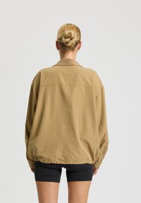Hellbraune übergroße Jacke mit glatter Textur, geripptem Kragen und elastischen Bündchen, kombiniert mit schwarzen Shorts.