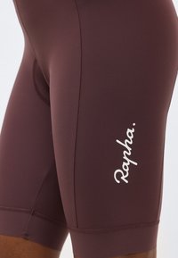 Gros plan sur un cuissard de cyclisme marron avec le logo blanc incurvé "Rapha" sur la cuisse, porté par une personne.