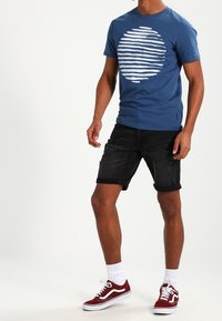 Camiseta de algodón azul con un gráfico en forma de círculo de rayas blancas horizontales, combinada con pantalones cortos de mezclilla negros con bordes deshilachados y zapatillas rojas.