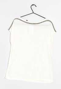 Túnica blanca de corte holgado con un escote redondo, textura suave y adornos metálicos en los hombros. Cuélgala de un gancho negro.