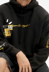 Felpa con cappuccio nera con disegni grafici gialli e bianchi, inclusi testo e toppe. Dotata di cappuccio con coulisse e polsini a coste. Maniche corte visibili.