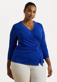 Blue wrap top made of smooth fabric, featuring a V-neckline and three-quarter sleeves. Tied at the waist with subtle draping detail.