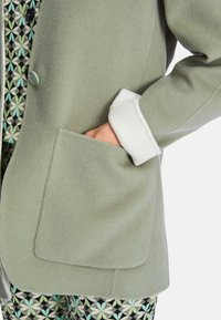 Grüne Wollmischjacke mit einer großen aufgesetzten Tasche und weißen Kunstfellmanschetten. Strukturiertes Material mit glattem Futter und subtilen Details.