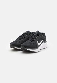 Schwarze Sportschuhe mit einem atmungsaktiven Mesh-Obermaterial, flachen Schnürsenkeln, gepolsterten Sohlen und einem weißen Logo an der Seite. Strukturierte Gummisohle.
