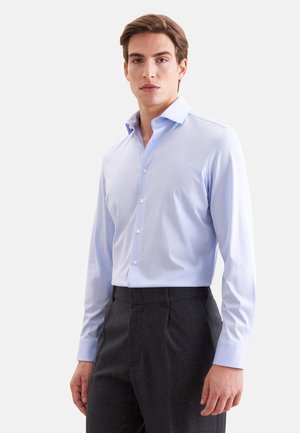 Jeune homme portant une chemise habillée bleu clair à manches longues rentrée dans un pantalon gris foncé, debout devant un fond clair uni.