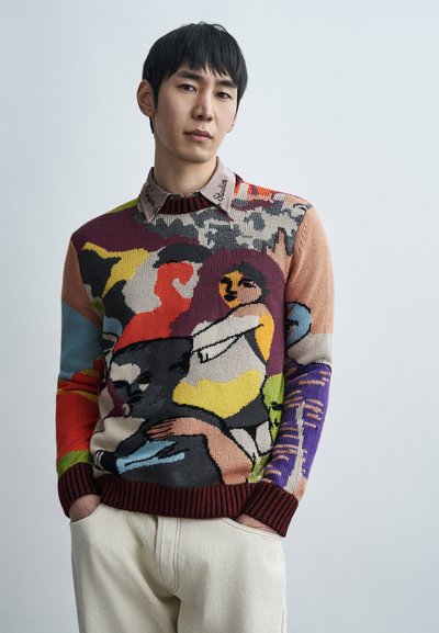Pull en tricot avec un motif abstrait multicolore incluant des figures. Le tissu est doux avec un ourlet côtelé bordeaux. Pantalon beige avec des poches.