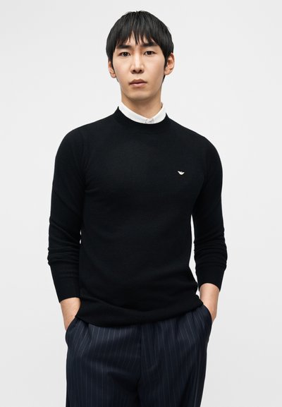 Μαύρο πλεκτό πουλόβερ με στρογγυλή λαιμόκοψη, που διαθέτει μικρό λογότυπο στο στήθος. Το μοντέλο φοράει πουκάμισο με γιακά από κάτω, συνδυασμένο με παντελόνι με ρίγες.