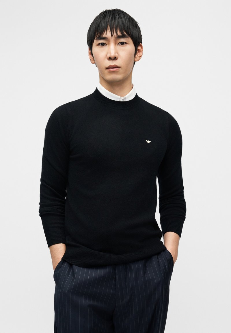 Pull noir en maille avec un col rond, orné d'un petit logo sur la poitrine. Le modèle porte une chemise à col en dessous, associée à un pantalon à fines rayures.