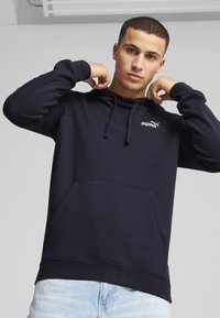 Námornícky modrá mikina s kapucňou vyrobená z bavlnenej zmesi, s prednou kapsou, kapucňou so šnúrkami a malým bielym logom Puma na hrudi. Mäkká textúra.