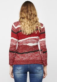 Rot-weißer gestrickter Pullover mit horizontalen Wellenmustern, schwarzen Akzenten, langen Ärmeln und einer taillierten Passform, kombiniert mit blauen Jeans.