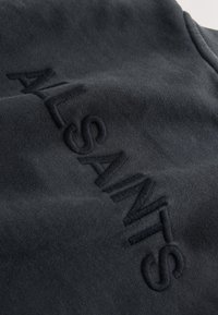 Schwarzer Stoff mit erhabenem, besticktem Text "ALSANDS" in diagonaler Anordnung.