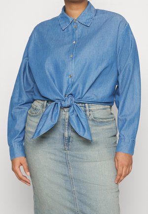 Un mannequin porte une chemise bleue boutonnée et nouée à la taille, avec une jupe en jean délavé clair.